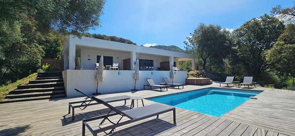Un espace piscine extérieur moderne, doté d’une terrasse en bois, de chaises longues et d’un bâtiment blanc avec un patio couvert.