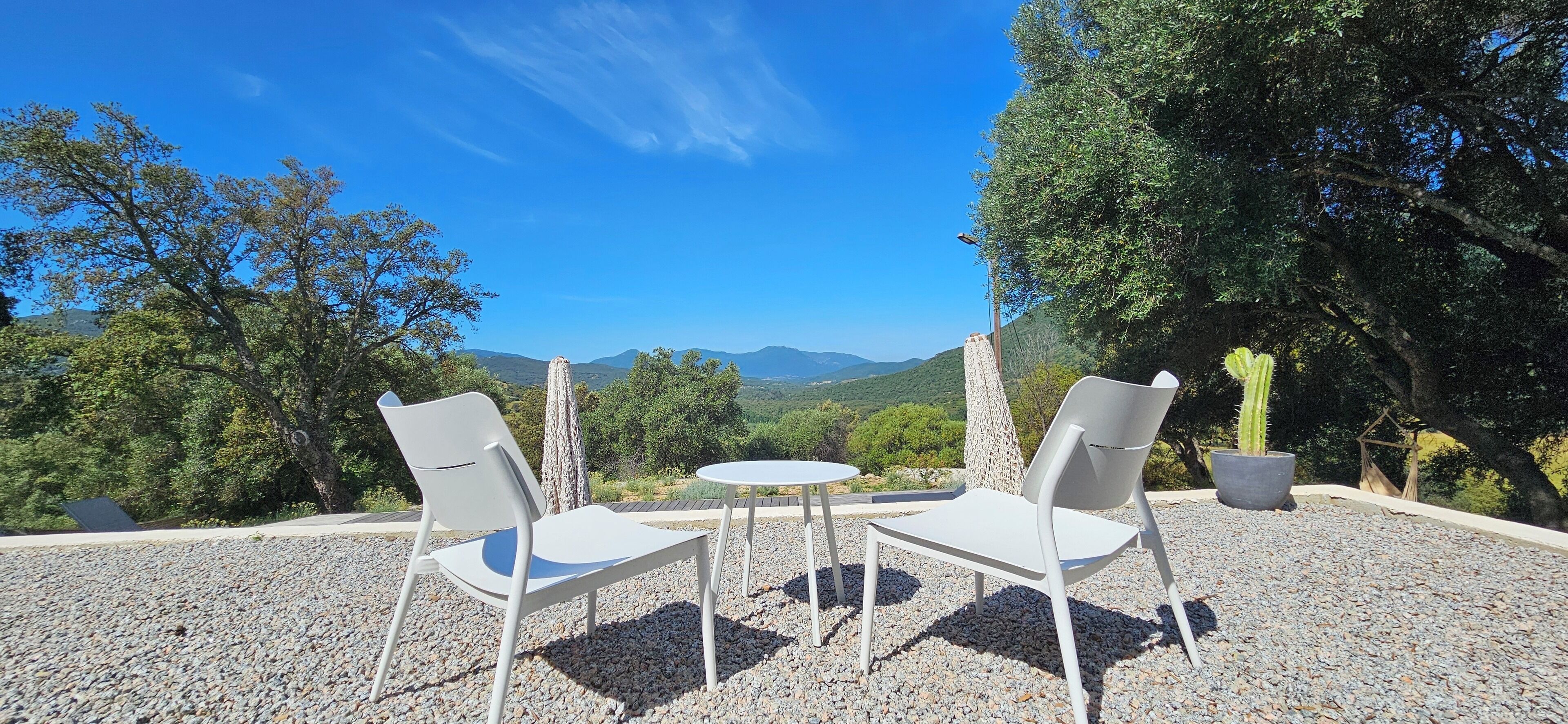 An outdoor seating area with two white chairs and a small round table, surrounded by gravel, with a view of green hills and a clear blue sky.