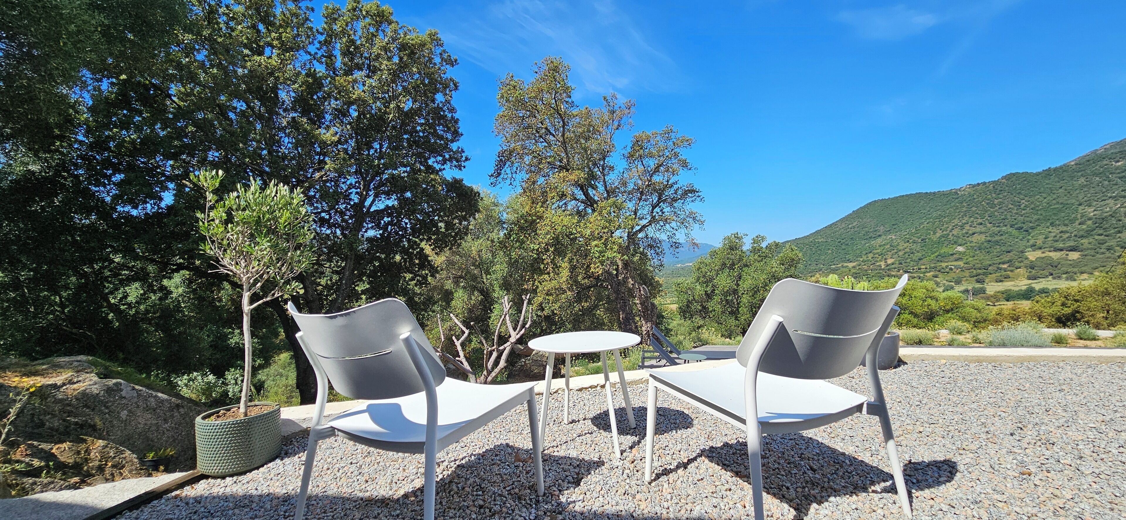 Two white chairs and a small round table set on a gravel surface, surrounded by trees and a mountainous backdrop.