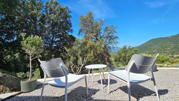Two white chairs and a small round table set on a gravel surface, surrounded by trees and a mountainous backdrop.