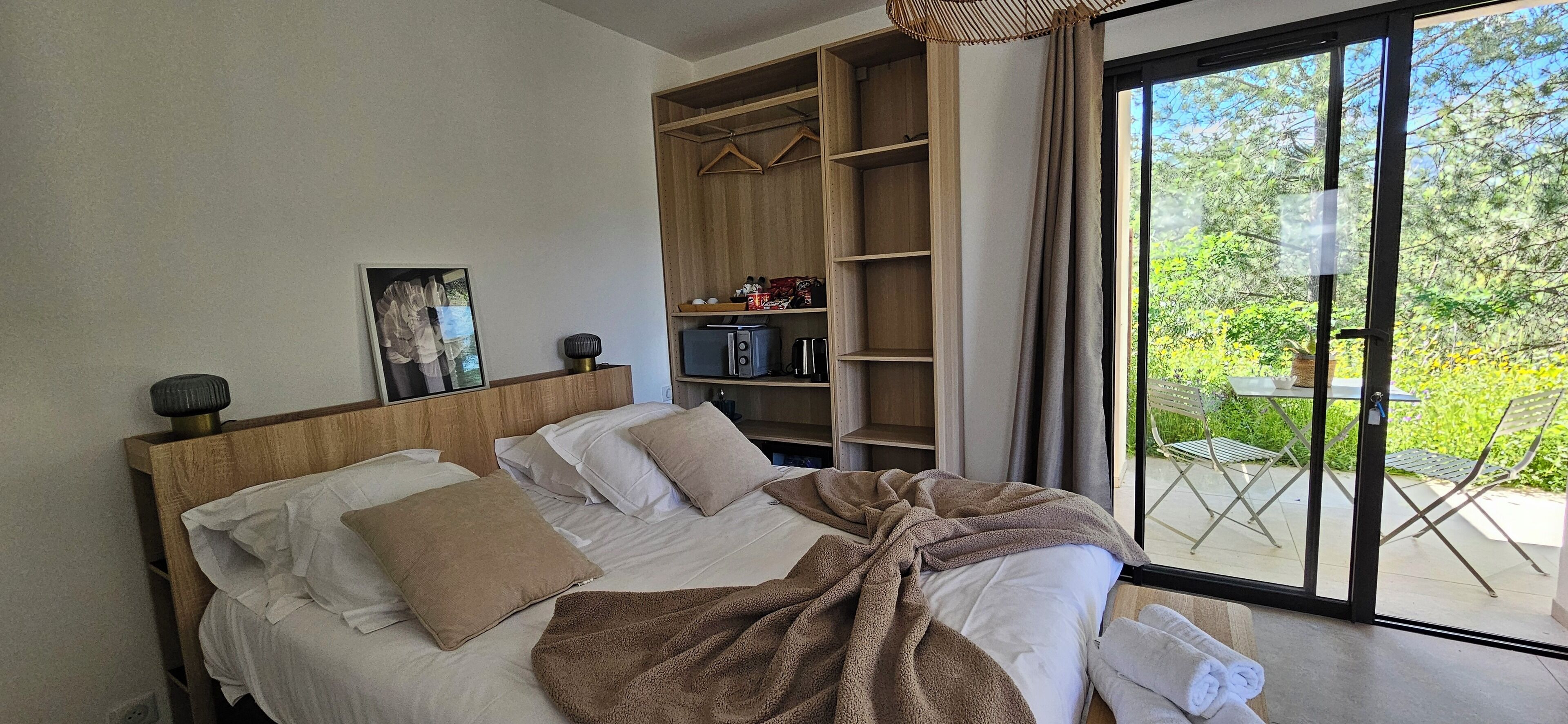 A bedroom with a large bed, a wooden wardrobe, and a view of a garden through a sliding glass door.