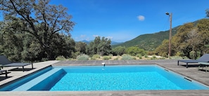 Ein rechteckiger Swimmingpool mit umlaufender Holzterrasse, auf jeder Seite jeweils zwei Liegestühle und im Hintergrund eine Berglandschaft.