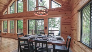 A wooden cabin interior with a dining table set for four, large windows, and a chandelier. - Make Memories In This Top Notch Lakefront Cabin (Stone Lake)
