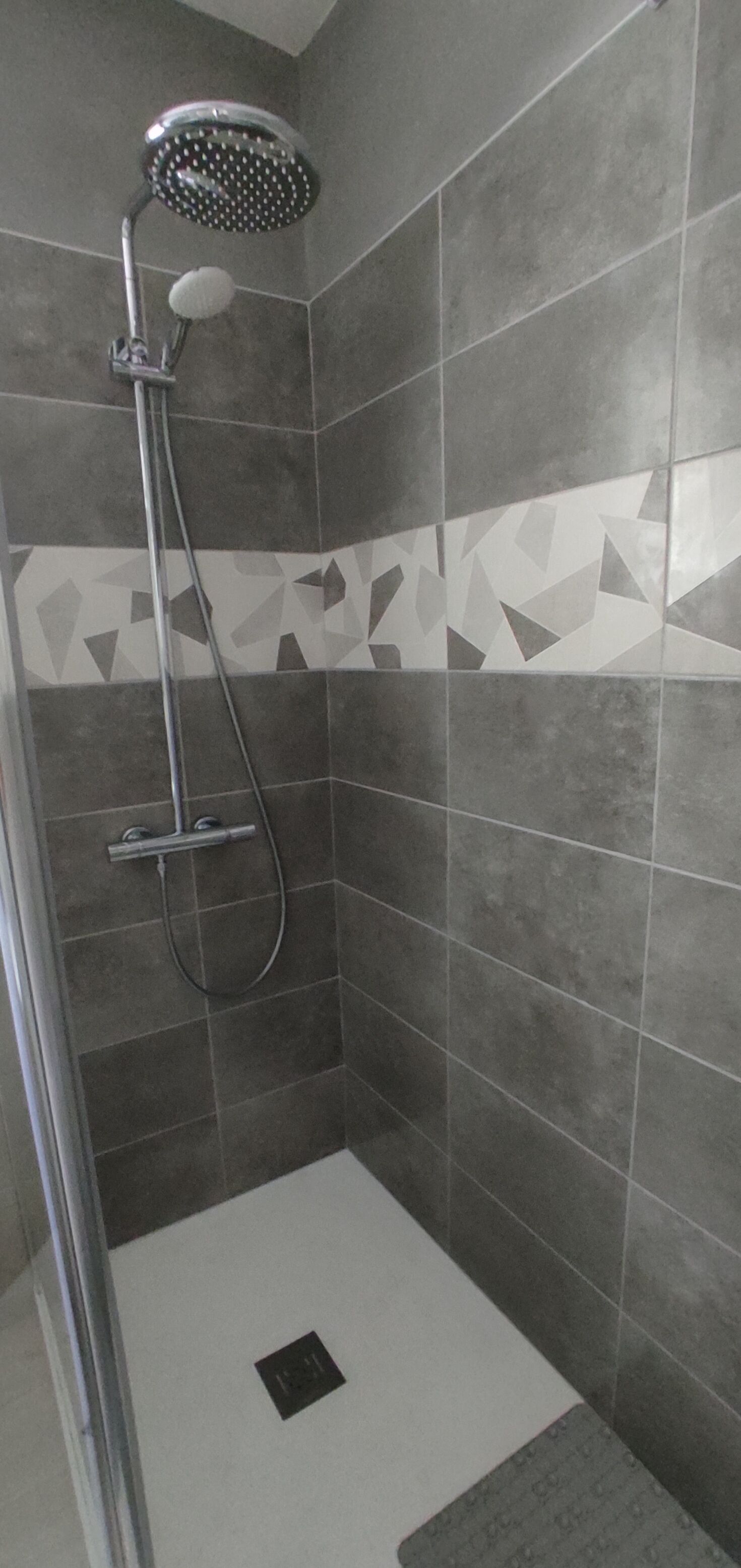 A shower with a handheld showerhead and a wall-mounted faucet. The shower is enclosed with grey tiles and has a geometric pattern on the lower wall.