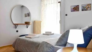 Un dormitorio con cama, mesita de noche con lámpara, espejo redondo, ventana con cortinas y cuadros en la pared.