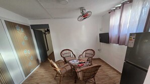 Una habitación pequeña y moderna con piso de madera, una mesa de comedor con cuatro sillas, un ventilador de techo, un refrigerador negro y un televisor montado en la pared.