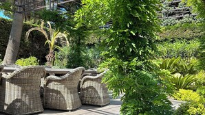 A wooden deck with wicker furniture, surrounded by lush greenery and flowering plants.