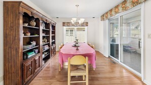 A dining area with a pink table, wooden chairs, a chandelier, and a large wooden cabinet with shelves.