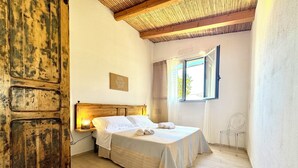 A bedroom with a wooden door, a bed with white linens, a bedside table with a lamp, and a window with curtains. - "Le Corti Sarde Sea Breeze" (Bari Sardo)