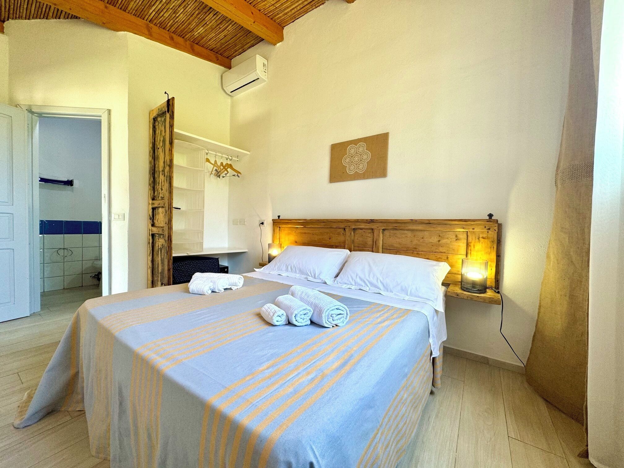 A bedroom with a bed, towels, a wooden headboard, an air conditioning unit, and a wall-mounted light fixture.