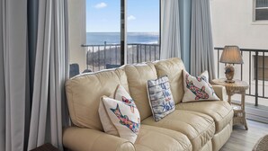 En beige lädersoffa med dekorativa kuddar, ett sidobord i rotting med en lampa och en utsikt över havet genom stora fönster.