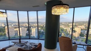 Una habitació àmplia amb un gran finestral que ofereix vistes a la ciutat, una làmpada moderna de cristall i una taula rodona amb cadires.