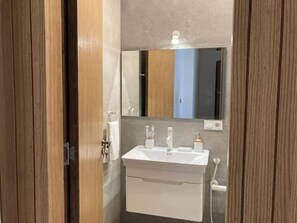 Une salle de bain avec une porte en bois, un lavabo blanc, un miroir et une serviette accrochée au mur.