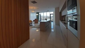 Una cuina moderna amb una illa gran, zona de menjador amb cadires i vista a la ciutat a través de grans finestres.