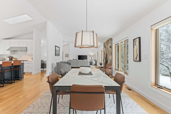A modern kitchen with white cabinets, a dining area with a marble table, and a large window with a view of trees.