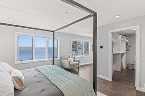 Quarto com cama de dossel, janelas amplas, vista para a água, uma cadeira e armário com roupas penduradas.