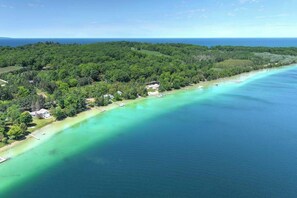 An aerial view of a coastal area with a clear turquoise sea, dense green forest, and a few buildings along the shoreline.