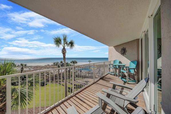 A balcony with a view of the beach and ocean, wooden deck, and outdoor furniture.
