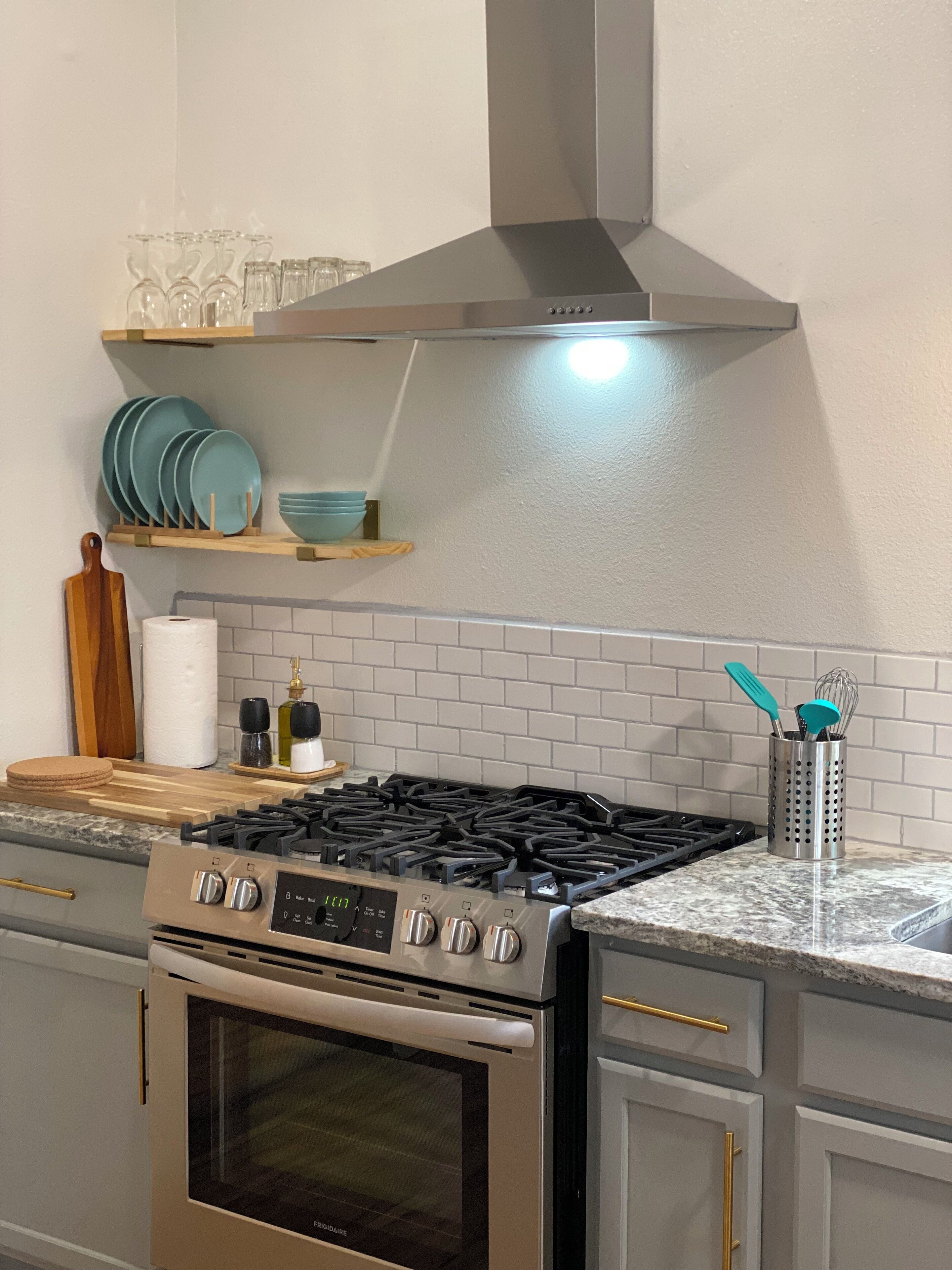 Une cuisine équipée d’une cuisinière, d’un four et d’une hotte en acier inoxydable, le tout placé au-dessus de la cuisinière. On y trouve des étagères remplies d’assiettes et de verres, ainsi qu’un porte-épices sur le plan de travail.