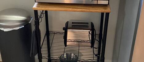 Una cocina con un microondas sobre una estantería de madera, un cubo de basura y una ventana con vistas a una valla.