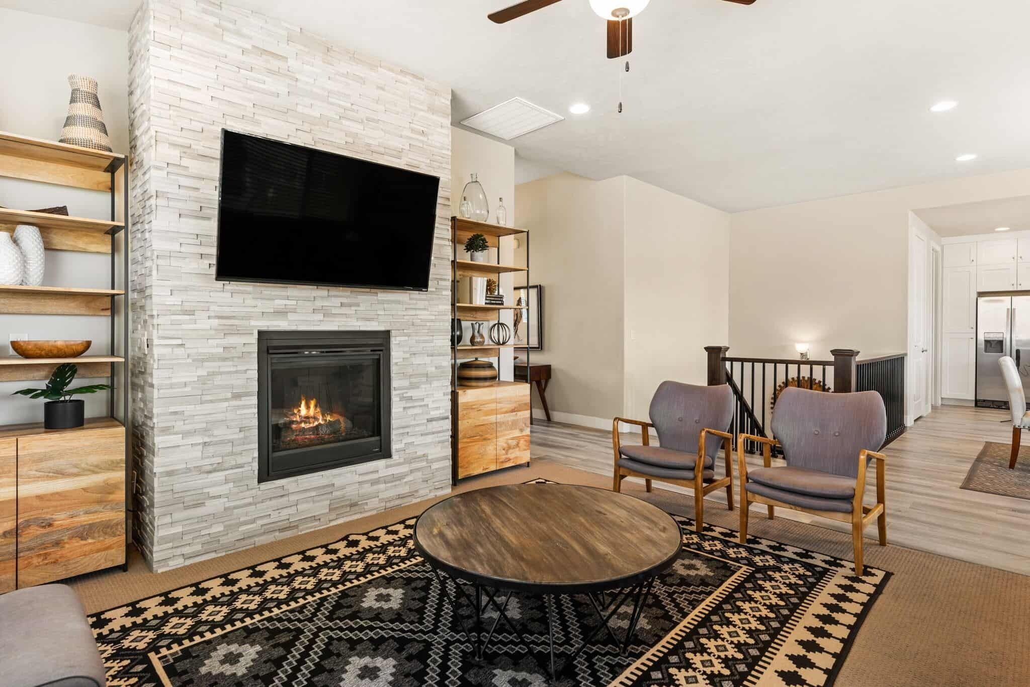 A modern living room with a fireplace, a flat-screen TV mounted above it, and a round coffee table. There are two armchairs and a bookshelf with decorative items.