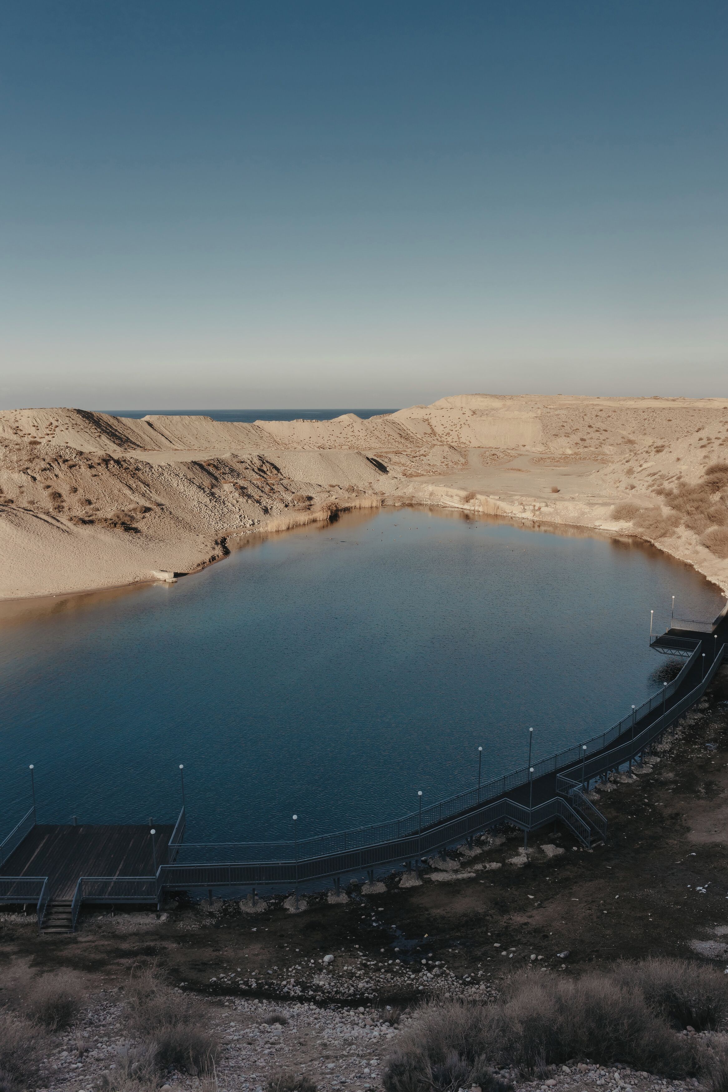 Un extenso cuerpo de agua circular rodeado de terreno árido, con un muelle de madera y escalones que bajan al agua.