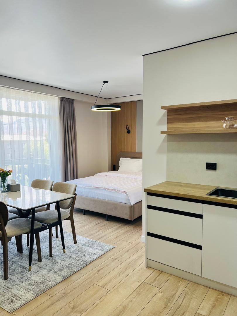 A modern hotel room with a dining table, chairs, a bed, a kitchenette, and a large window with curtains.