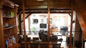 Un intérieur rustique avec des poutres en bois, un coin repas comprenant des chaises et une table, un canapé, un téléviseur et une bibliothèque.