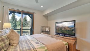 A bedroom with a bed, a television, a window with a view of trees, and a ceiling fan.