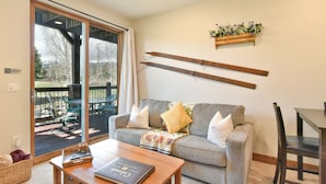A cozy living room with a grey sofa, a wooden coffee table, and a dining table with chairs. A sliding glass door leads to an outdoor deck with chairs.