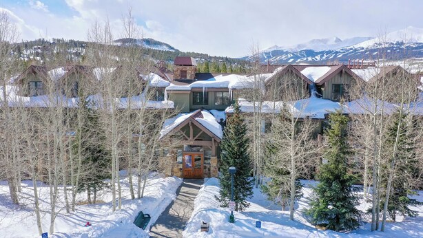 A snow-covered lodge with a central building surrounded by trees, set against a backdrop of mountains.