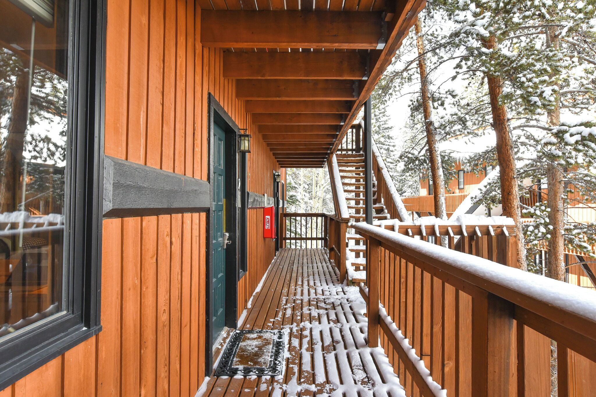 Una terraza de madera con barandilla metálica, una puerta y un pequeño espacio al aire libre cubierto de nieve.