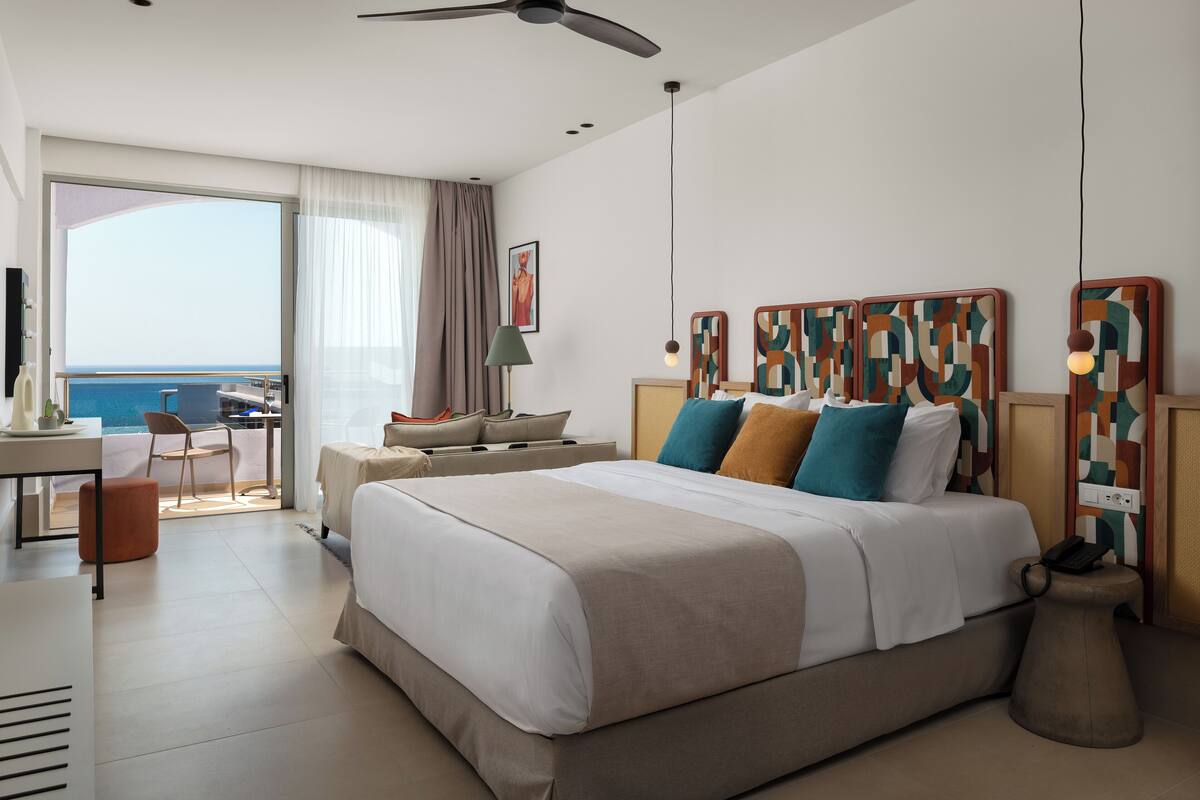 A modern hotel room with a large bed, a desk, a chair, and a view of the sea.