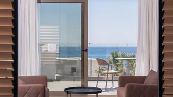 A room with a sliding glass door leading to a balcony with a view of the sea. There are two chairs and a small table on the balcony.