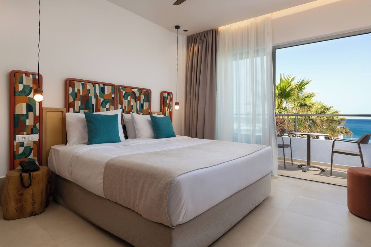 A modern bedroom with a large bed, a bedside table with a lamp, and a view of the ocean through a balcony.