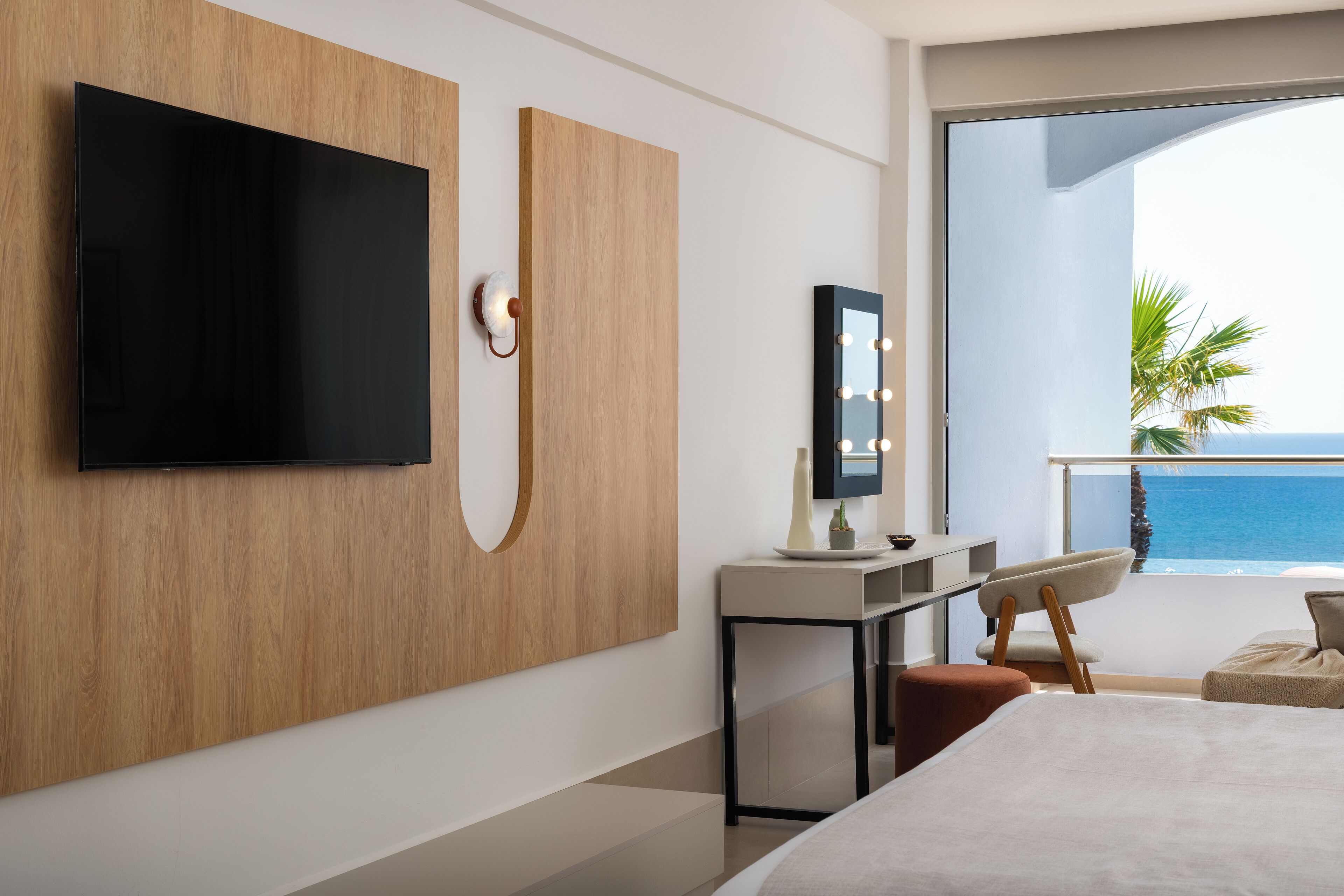 A modern hotel room with a large bed, a flat-screen TV mounted on a wooden panel, a vanity with a mirror and lights, and a balcony with a view of the ocean.