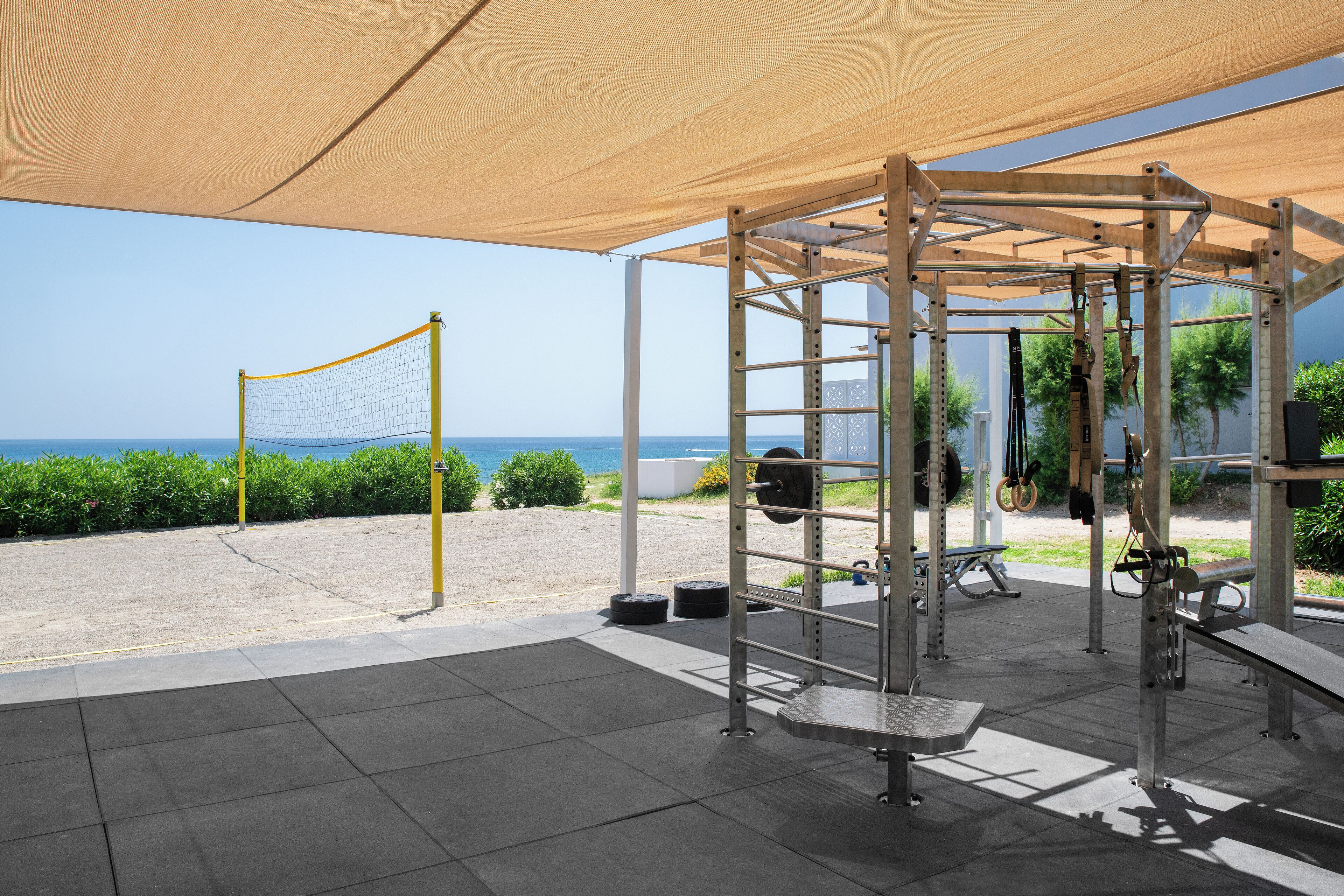 Un gimnasio al aire libre con una cancha de voleibol cubierta por una red, equipo de levantamiento de pesas y vista al mar.