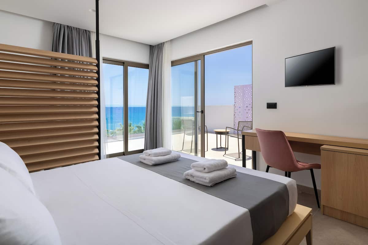 A hotel room with a bed, a desk, a chair, a TV, and a balcony with a view of the sea.