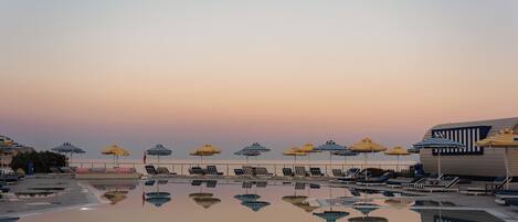 Un espace aménagé au bord de la piscine, avec des chaises longues et des parasols, dont le reflet se mire dans l’eau.