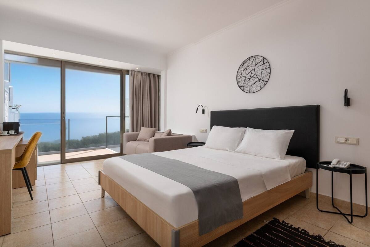 A modern hotel room with a large bed, a desk, and a chair. There is a view of the sea through the large window.