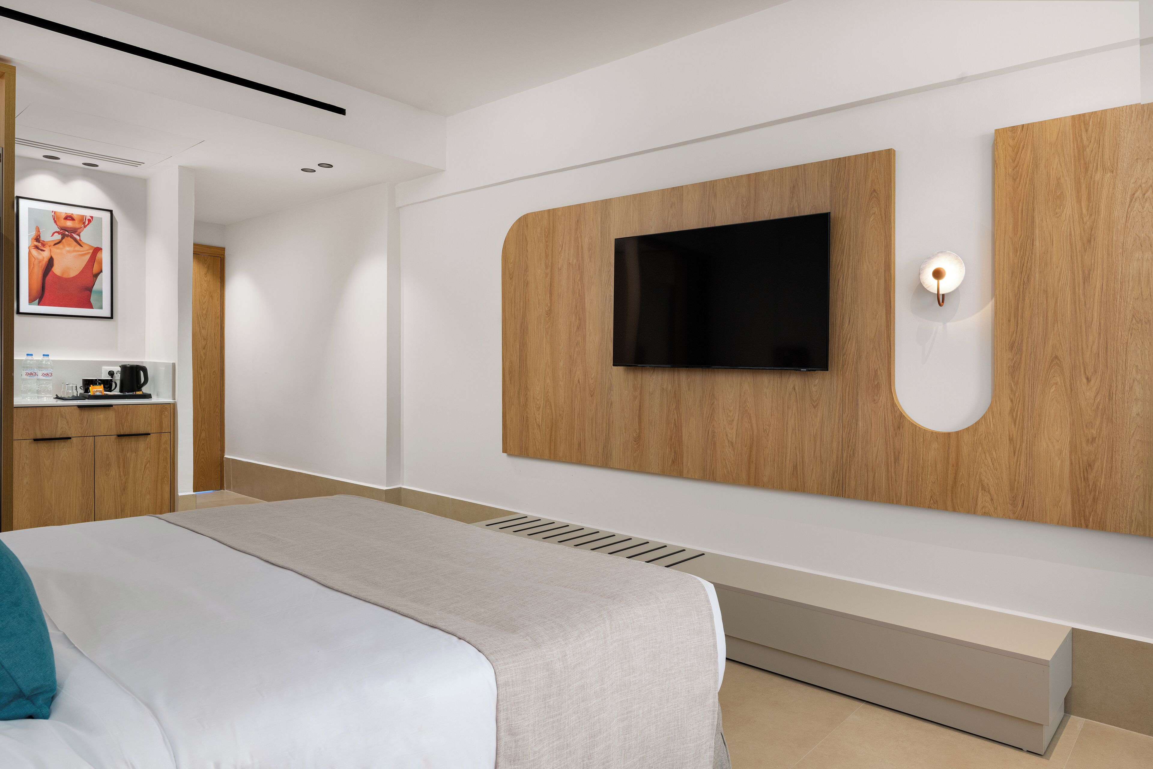 A modern hotel room with a large bed, a flat-screen TV mounted on a wooden panel, a small bedside table with a lamp, and a framed picture on the wall.
