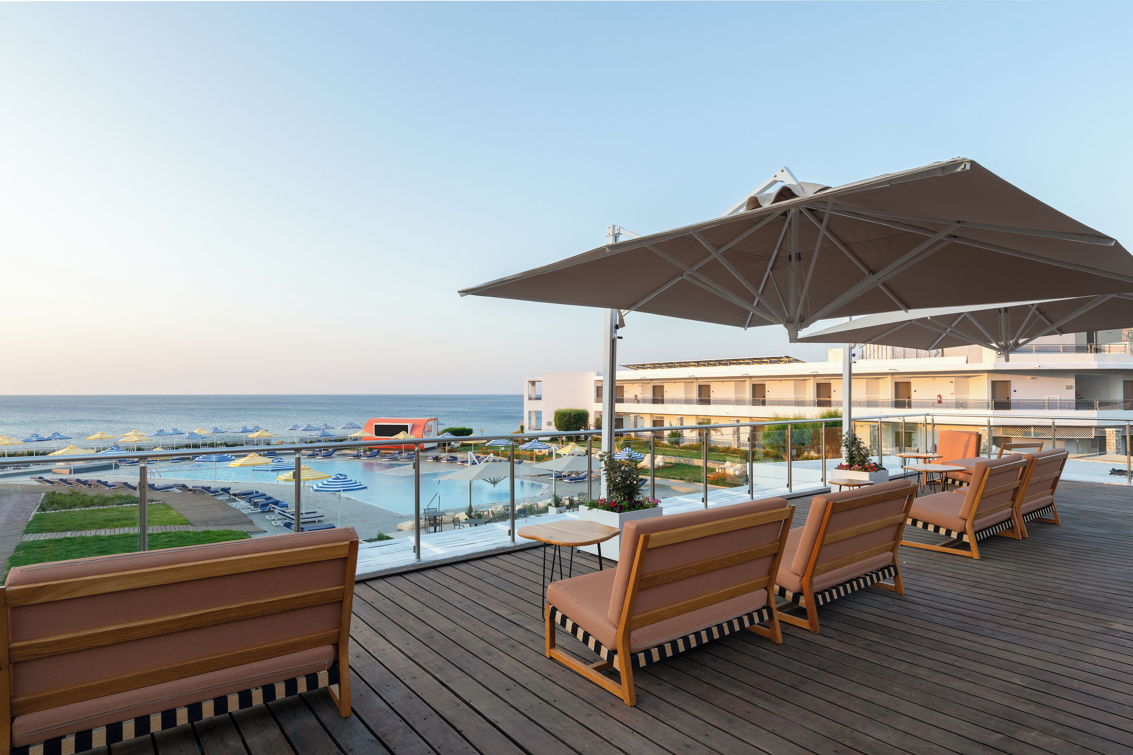 A wooden deck with outdoor seating and a large umbrella, overlooking a pool and beach area.