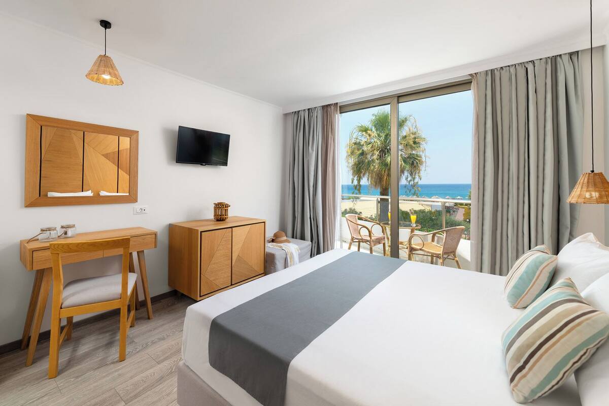 A hotel room with a bed, desk, and chair, a TV, and a balcony with a view of the beach and ocean.
