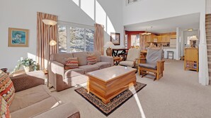 A spacious living area with a sofa, armchair, and coffee table, a kitchen in the background, and large windows allowing natural light.