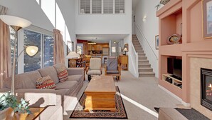 A spacious living area with a high ceiling, a fireplace, a staircase, and a kitchen in the background.