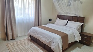 A neatly made bed with a wooden headboard, a bedside table with drawers, and a window with sheer curtains. - Oreville Apartments Namugongo Seeta Rd (Seeta)