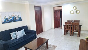 A living room with a dark blue sofa, a wooden coffee table, and a dining area with a wooden table and chairs. - Oreville Apartments Namugongo Seeta Rd (Seeta)