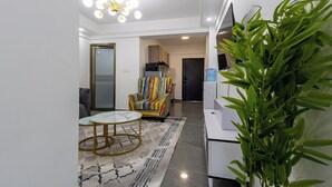 A modern living room with a glass coffee table, a colorful armchair, a potted plant, and a TV on a white cabinet. - Erica Suites - Riverside 1BR Apartments (Nairobi)