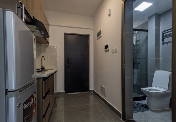 A compact kitchen with a refrigerator, stove, and microwave. A door leads to a bathroom with a toilet and shower.