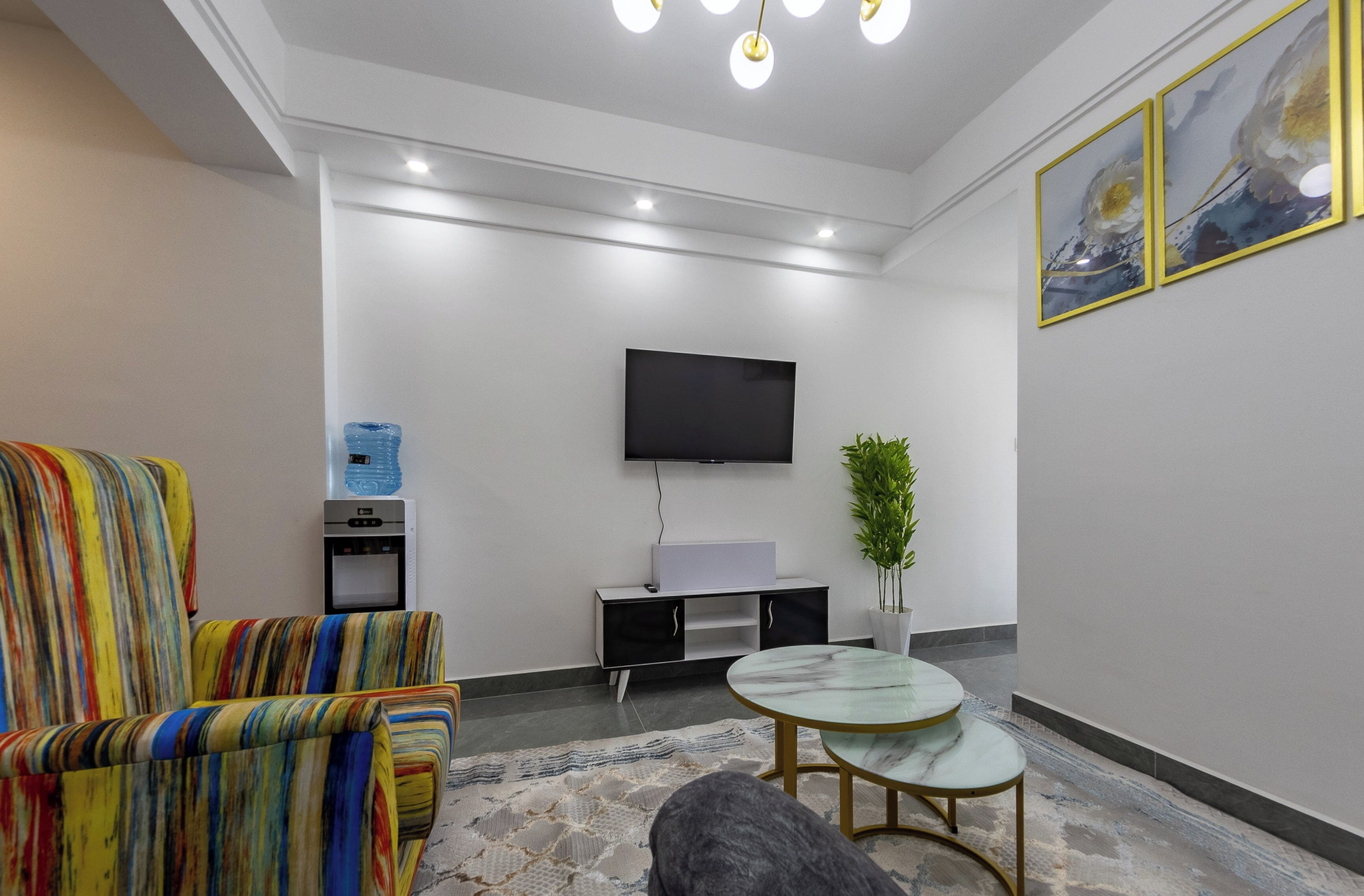 A modern living room with a flat-screen TV mounted on the wall, a striped armchair, a small round table, and a water dispenser.